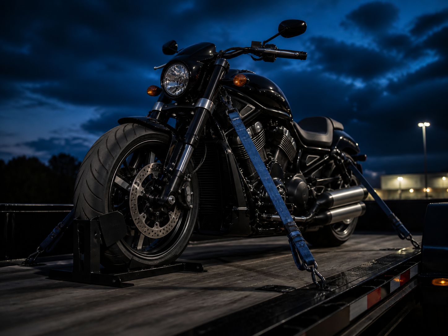 Black cruiser motorcycle secured with blue straps and a wheel chock on a trailer deck at dusk
